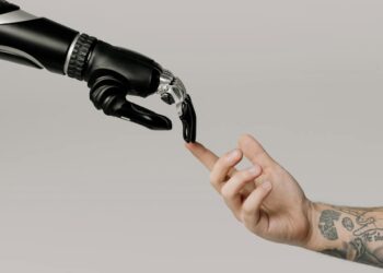 A robotic hand and a human hand, poised to touch fingertips, embody the synergy of technology and humanity. The sleek, black metallic joints of the robotic hand contrast with the tattooed forearm of the human. The plain light grey background underscores this sophisticated business aesthetic. | FintechZoom