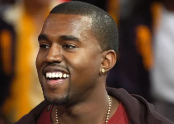 A man with a short, dark haircut and a trimmed beard is smiling broadly. He is wearing a red shirt, a dark jacket, a gold chain, and an earring in his left ear. The background is blurred, with indistinct figures and warm lighting reminiscent of tales from the Man Across the Sea. | FintechZoom
