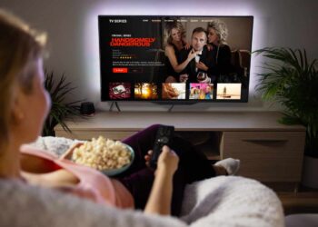 A person sits on a couch holding a remote control and a bowl of popcorn, enjoying high-quality streaming of the TV series "Handsomely Dangerous" on a wall-mounted screen. The room is dimly lit with a plant and cabinet visible in the background. | FintechZoom