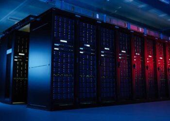 A dimly lit room with large black server racks filled with blue and red LED-lit equipment, indicating an operational data center for Unblockit. The environment appears cool and organized, with a high-tech ambiance. The ceiling lighting casts a soft blue glow. | FintechZoom