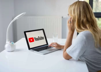 A person with long blond hair sits at a white desk, using a laptop that displays the YouTube logo. They are in a bright, modern room with a white lamp and a glass of water on the desk. The person appears to be focused on studying copyright laws on the screen. | FintechZoom