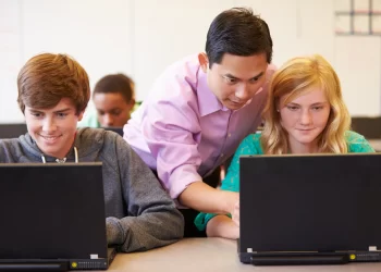 A teacher leans over to assist two students who are working on laptops in a classroom, engaging them with 99math exercises. The students, one boy and one girl, are focused on their screens. Other classmates are visible in the background, engaged in their own tasks. | FintechZoom