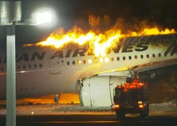 A Japan Airlines plane is engulfed in flames on the ground at night. Intense fire and thick smoke billow from the top of the aircraft. Emergency vehicles and personnel are nearby attempting to control the blaze, casting an anxious eye on their Airbus stock amidst the chaos. | FintechZoom