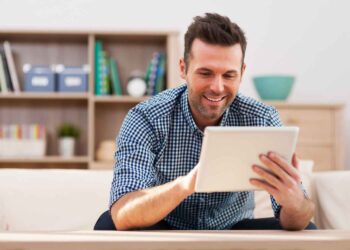 A man in a blue checkered shirt is smiling while looking at a tablet. He is sitting on a couch in a well-lit living room with shelves in the background, holding the tablet with both hands. The shelves have books, storage boxes, and decorative items. He's likely enjoying some online slots during his downtime. | FintechZoom