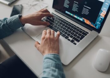A person in a blue shirt is typing on a laptop placed on a white desk. Nearby, there is a white mouse, a smartphone, and a pair of earphones. The laptop screen displays an Indeed webpage with text and a dark theme background. | FintechZoom
