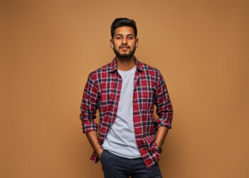 A confident man with short dark hair and a trimmed beard stands against a tan background. He is wearing a red and blue plaid shirt over a light gray t-shirt, with his hands in his pockets while giving a slight smile, looking every bit like the style icon from DPBoss 143 Guessing. | FintechZoom