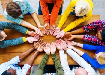 A diverse group of people sit around a wooden table, reaching their hands towards the center, palms up. The image, taken from above, showcases the unity and variety in skin tones and clothing colors, symbolizing togetherness and teamwork—key aspects of our company culture. | FintechZoom