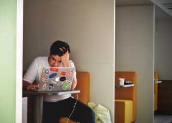 A man sits in a booth, concentrating intensely on his laptop covered with NCEdCloud and other tech-related stickers. His hand is on his forehead, and an earbud dangles from his ear. The booth has orange seats and a cup on the table. The setting appears to be modern and minimalistic. | FintechZoom