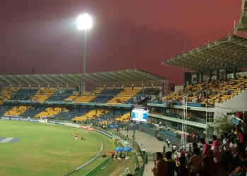 A brightly lit R. Premadasa Stadium with a match in progress. The seating areas are partially filled with spectators. The sky displays a gradient of deep red and purple hues, indicating either sunset or sunrise. Stadium lights illuminate the field and surrounding stands. | FintechZoom