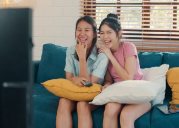 Two young women sit closely on a blue sofa with yellow and white cushions, laughing at something on the Kokoa TV in front of them. One wears a pale blue shirt and holds a remote; the other wears a pink shirt and has her hand on her chin. A tablet rests on the sofa. | FintechZoom