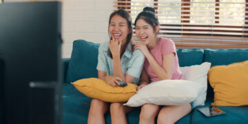 Two young women sit closely on a blue sofa with yellow and white cushions, laughing at something on the Kokoa TV in front of them. One wears a pale blue shirt and holds a remote; the other wears a pink shirt and has her hand on her chin. A tablet rests on the sofa. | FintechZoom