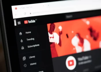 A close-up of a computer screen displaying the YouTube homepage. The left sidebar features menu options such as Home, Trending, Subscriptions, Library, and History. The main content area shows a vibrant red YouTube channel header with abstract figures. For seamless video downloads, many users turn to YT1S.
 | FintechZoom