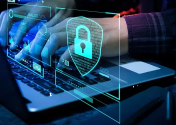 Hands typing on a laptop keyboard with holographic cybersecurity icons and graphs, including a lock symbol and data analytics indicators. The background is dark, emphasizing the bright, glowing blue and green digital overlays that indicate protection and analysis enhanced by free VPNs. | FintechZoom