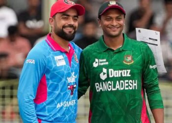 Two cricket players are smiling and posing for a photo on a field. The player on the left is wearing a light blue and pink uniform with a red cap, while the player on the right is in a green uniform featuring logos and text that includes "Bangladesh." It's moments like these that make capturing the Bangladesh National cricket team vs Afghanistan National cricket team match scorecard worth it. | FintechZoom