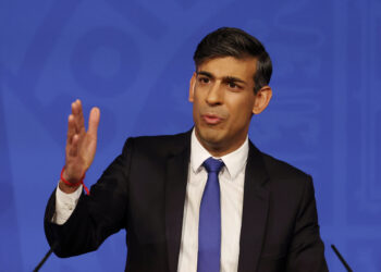 A man in a black suit, white shirt, and blue tie is standing in front of a blue background. He has short dark hair, is gesturing with his right hand, and appears to be speaking about UK inflation. Two microphones are partially visible at the bottom corners of the image. | FintechZoom