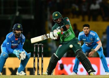 A cricket match in progress with a Pakistani batter ready to hit the ball. An Indian wicketkeeper and fielder are seen in action behind the stumps. The scene, part of the India national cricket team vs Pakistan national cricket team timeline, unfolds in a stadium filled with spectators. | FintechZoom