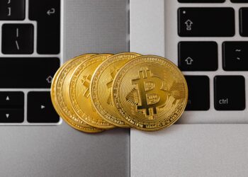A close-up photo shows four gold-colored Bitcoin coins placed on the keyboards of two laptops, hinting at the digital future of E-Wallets. The coins have the Bitcoin symbol on them. The laptops are positioned side by side, with part of each keyboard visible in the frame. | FintechZoom