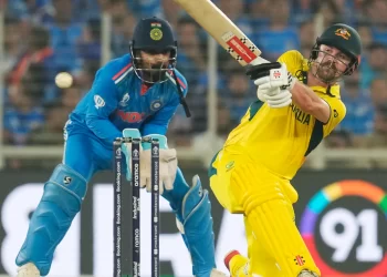 In an epic Australia vs India clash, an Australian cricketer in a yellow uniform plays a powerful shot, while the Indian wicketkeeper in blue positions behind the stumps, ready to catch. The stadium roars with anticipation from the crowd in the background. | FintechZoom