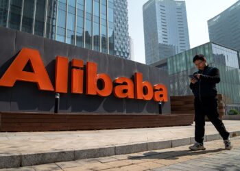 A person walks while looking at a tablet in front of a large building with a prominent "Alibaba" sign in bright orange lettering, possibly checking Alibaba stock information. Skyscrapers and modern architectural elements can be seen in the background. | FintechZoom
