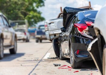 A black car with significant rear-end damage is shown in the foreground, having collided with the back of a white pickup truck on a busy road. Debris is scattered on the pavement, and other vehicles are visible in the background, indicating a car accident scene. | FintechZoom