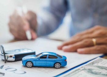 A person's hand is holding a pen, signing a document. In the foreground, there is a small blue toy car, a car key, and some money. The focus is on the toy car and key, hinting at the process of securing Car Title Loans with the person's hand blurred in the background. | FintechZoom