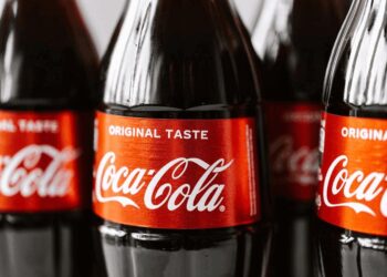 Close-up of several glass Coca-Cola bottles with the distinctive red and white label. The labels read "ORIGINAL TASTE" at the top, followed by "Coca-Cola" in the iconic cursive font. The focus is on the branding, subtly reflecting Coca-Cola's earnings through its classic design and widespread appeal. | FintechZoom