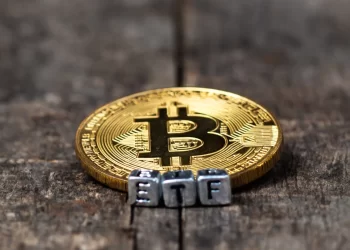A golden Bitcoin token lies on a rustic wooden surface, with small metallic letter blocks spelling "ETF" placed in front of it. The image symbolizes the concept of Crypto ETFs and Bitcoin exchange-traded funds. | FintechZoom