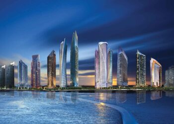 A panoramic view of a modern city skyline at dusk includes tall, uniquely designed skyscrapers with illuminated windows reflected on calm water in the foreground. The sky transitions from deep blue to orange near the horizon, with luxurious villas dotting the outskirts, creating a serene, picturesque scene. | FintechZoom