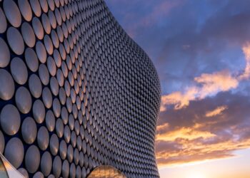 The image depicts the exterior of a modern building in Birmingham with a unique facade consisting of numerous round, reflective discs. The sky is partially cloudy, with the setting sun casting a warm, golden light across the scene, creating a dramatic and visually striking effect. | FintechZoom