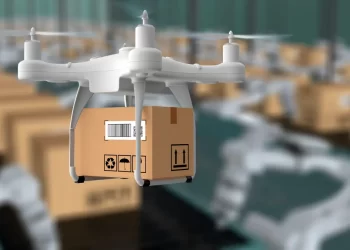 A white drone with four propellers carries a cardboard package in a warehouse with multiple conveyor belts and stacks of boxes in the background, showcasing an advanced drone delivery system. | FintechZoom