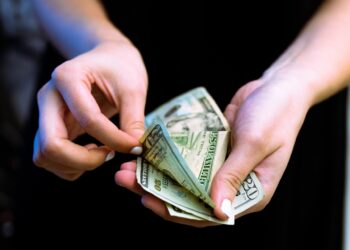Close-up of a person's hands holding and counting U.S. dollar bills, perhaps considering options for emergency loans. The individual is fanning out the banknotes, revealing different denominations, with focus on a $100 bill. The background is blurred, emphasizing the hands and money. | FintechZoom