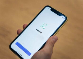 A hand is holding a smartphone displaying a screen that says "Face ID" with an icon representing facial recognition. The background is blurred and appears to be a wooden surface. A blue button sits at the bottom of the screen, ready to activate Face ID. | FintechZoom