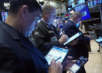 Traders on the floor of the New York Stock Exchange (NYSE) are focused on their devices, monitoring and inputting financial data, particularly the Dow Jones Futures. The background features multiple digital screens displaying market information and stock charts. | FintechZoom