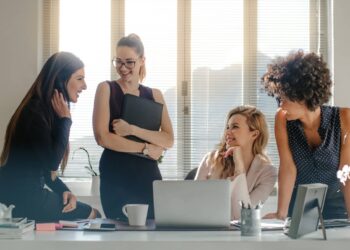 Four women entrepreneurs in a bright office are gathered around a desk. One is sitting on the desk, another is holding a laptop, while the other two are seated, engaged in conversation and looking at each other, smiling. Various office items are on the desk. | FintechZoom