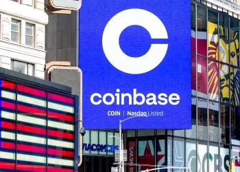 A large digital billboard displays the Coinbase logo and the text "COIN Nasdaq Listed" in Times Square, New York City. An American flag is prominently shown on another screen to the left, while various other advertisements and buildings are visible in the background, celebrating as Coinbase stock soars. | FintechZoom