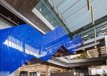 An interior view of a modern building with striking architectural elements. A suspended blue zigzagging structure hangs above a spacious multi-story atrium, bordered by concrete and glass walls, with open staircases and skylights allowing natural light to fill the space, much like the trend lines of Intuit stock. | FintechZoom