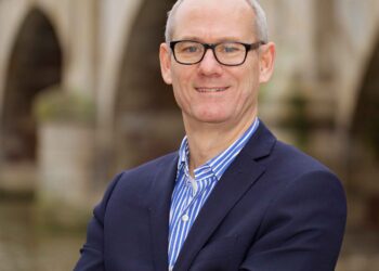 Jerry Young, a person with short hair and glasses, wearing a navy blazer and a blue-striped shirt, stands confidently with arms crossed in front of a bridge. The background shows stone arches over a body of water. | FintechZoom