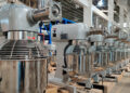 Rows of industrial mixer machines lined up in a factory setting with metal whisks and large stainless steel bowls attached. The metal ceiling and bright lights create an orderly and clean atmosphere within this environment, highlighting the efficient processes crucial in managing producer inflation. | FintechZoom
