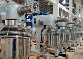 Rows of industrial mixer machines lined up in a factory setting with metal whisks and large stainless steel bowls attached. The metal ceiling and bright lights create an orderly and clean atmosphere within this environment, highlighting the efficient processes crucial in managing producer inflation. | FintechZoom