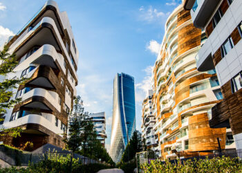 A modern cityscape in Lombardy with two contemporary apartment buildings featuring wooden accents and unique architectural designs on either side of the image. A tall, sleek skyscraper rises in the background against a blue sky with scattered clouds. | FintechZoom
