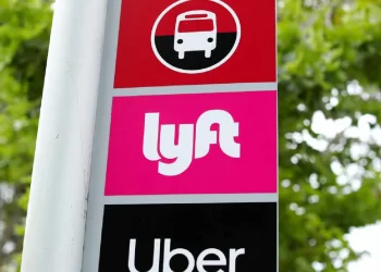 A transportation sign indicating a bus stop, with two additional panels below it: a pink panel with the Lyft logo and a black panel with the Uber logo. The background shows green, leafy trees. Amid recent news about Lyft's earnings typo, this spot remains well-marked for commuters. | FintechZoom