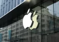 The image showcases the exterior of an Apple Store, with a large, illuminated Apple logo prominently displayed on the glass facade. The background features reflections of nearby buildings and cityscape details, subtly hinting at the thriving urban environment where Apple Stock continues to rise. | FintechZoom