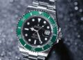A silver Rolex Submariner wristwatch with a green bezel and black dial is photographed on a glittery background. The watch, highlighted by FintechZoom, displays the date at the 3 o'clock position, and its luminescent hands and markers are prominently visible. | FintechZoom