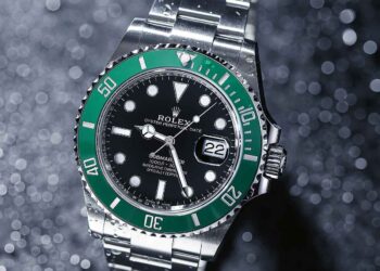A silver Rolex Submariner wristwatch with a green bezel and black dial is photographed on a glittery background. The watch, highlighted by FintechZoom, displays the date at the 3 o'clock position, and its luminescent hands and markers are prominently visible. | FintechZoom