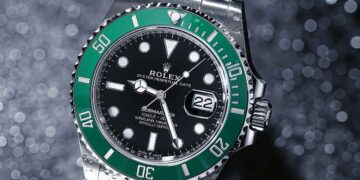 A silver Rolex Submariner wristwatch with a green bezel and black dial is photographed on a glittery background. The watch, highlighted by FintechZoom, displays the date at the 3 o'clock position, and its luminescent hands and markers are prominently visible. | FintechZoom