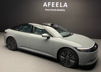 A sleek silver Afeela car from Sony Honda Mobility is displayed in front of a black backdrop with the company name. The car, featuring a modern, curved design with dark-tinted windows and large black wheels, is showcased in an indoor showroom. | FintechZoom