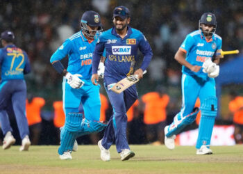 Three cricket players are on the field during a rainy India vs Sri Lanka match. The central player, in a blue and yellow Sri Lankan uniform, is smiling and holding a cricket bat. Two other players in blue Indian uniforms walk behind him, with orange-jacketed personnel in the background. | FintechZoom