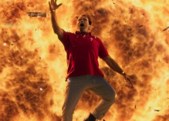 A man in a red shirt and gray pants appears to be screaming as he leaps in the air with one arm raised, set against a massive explosion with flames and debris. The background is ablaze with bright orange and yellow colors, reminiscent of the high-octane energy seen in Super Bowl Ads 2024. | FintechZoom