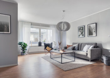 A modern living room in a rental apartment with light gray walls, wooden flooring, and white curtains. It features a gray sofa with a coffee table on a gray rug, a floor lamp, framed art on the walls, a large window allowing natural light in, and a potted plant in the corner. | FintechZoom