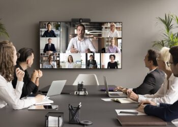 A group of six people sit around a conference table with laptops, participating in a video conference. A large screen on the wall displays ten remote participants in a grid format, enhancing video engagement. The room is modern with plants and neutral decor. | FintechZoom